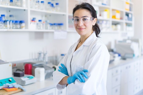A photo of smiling female chemist by shelves. Portrait of confident scientist standing with arms crossed. She is wearing protective eyeglasses and lab coat at laboratory.